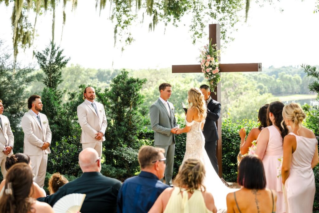 Emily and Aaron exchange vows during their outdoor Lakeview Gardens wedding ceremony with a wooden cross behind them.