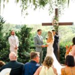 Emily and Aaron exchange vows during their outdoor Lakeview Gardens wedding ceremony with a wooden cross behind them.