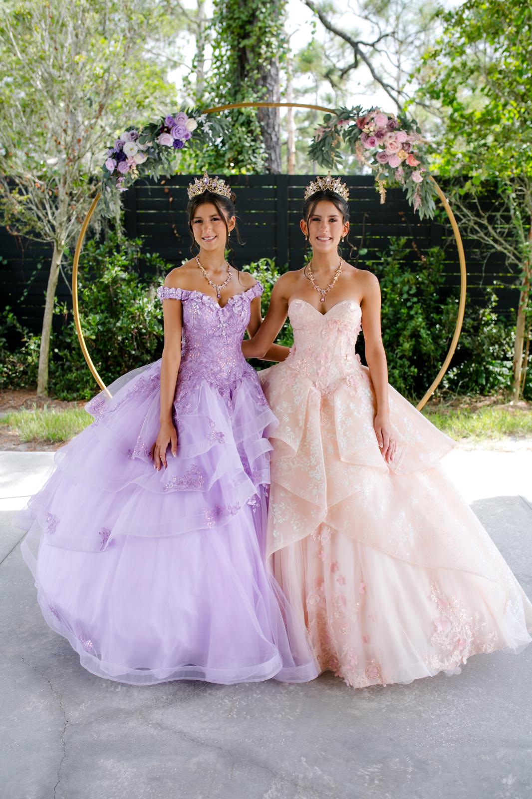 Alaina and London posing together in lavender and blush quinceañera gowns beneath a floral backdrop.